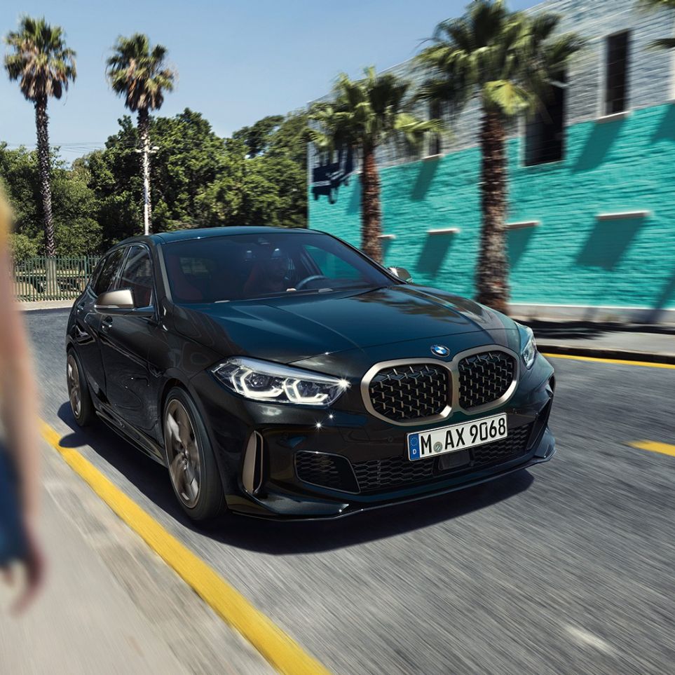 BMW M135i xDrive, три четвърти изглед отпред BMW M135i xDrive в Black Sapphire metallic, три четвърти изглед отпред в движение с жена на преден план
