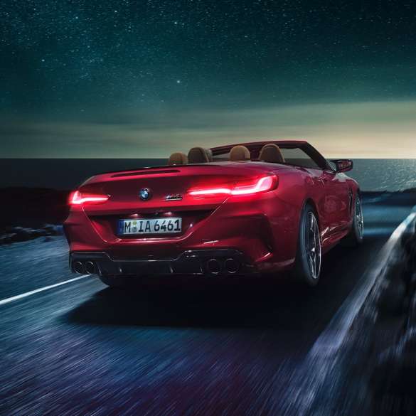 BMW M8 Competition Кабриолет в цвят Motegi Red metallic, три четвърти изглед отпред. BMW M8 Competition Кабриолет в цвят Motegi Red metallic с отворен мек покрив, три четвърти изглед отпред, в движение през нощта по крайбрежен път.