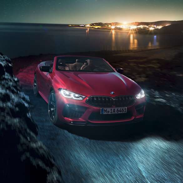 BMW M8 Competition Кабриолет в цвят Motegi Red metallic, три четвърти изглед отпред. BMW M8 Competition Кабриолет в цвят Motegi Red metallic с отворен мек покрив, три четвърти изглед отпред, в движение през нощта по крайбрежен път.