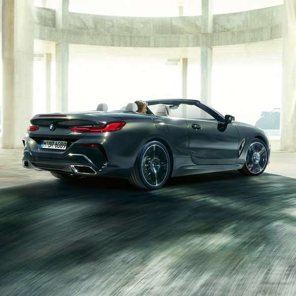 BMW M850i xDrive Кабриолет, три четвърти изглед отзад. BMW M850i xDrive Кабриолет в цвят Dravit Grey metallic с отворен мек покрив, три четвърти изглед отзад, в движение през бетонна сграда.