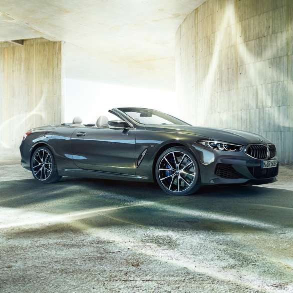 BMW M850i xDrive Кабриолет, три четвърти изглед отстрани. BMW M850i xDrive Кабриолет в цвят Dravit Grey metallic с отворен мек покрив, три четвърти изглед отстрани, в покой пред бетонна сграда.