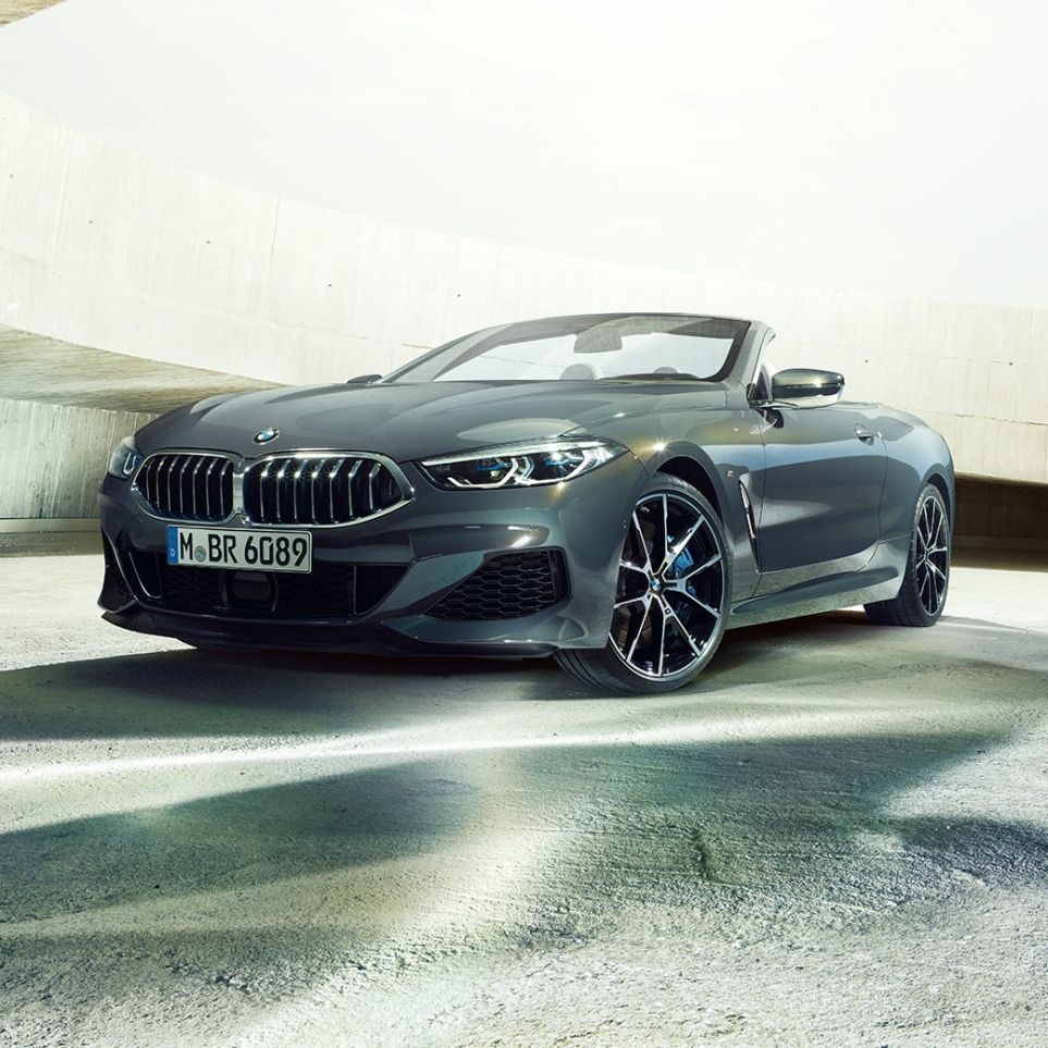BMW M850i xDrive Кабриолет, три четвърти изглед отпред. BMW M850i xDrive Кабриолет в цвят Dravit Grey metallic с отворен мек покрив, три четвърти изглед отпред, в покой пред бетонна сграда.