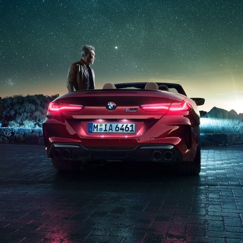 BMW M8 Competition Кабриолет, изглед отзад. BMW M8 Competition Кабриолет в цвят Motegi Red metallic с отворен мек покрив, изглед отзад, мъж стои до автомобила под звездно небе.