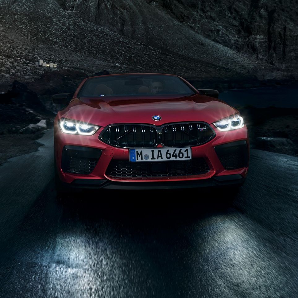 BMW M8 Competition Кабриолет в цвят Motegi Red metallic, изглед отпред. BMW M8 Competition Кабриолет в цвят Motegi Red metallic със светещи фарове, изглед отпред в движение през нощта.