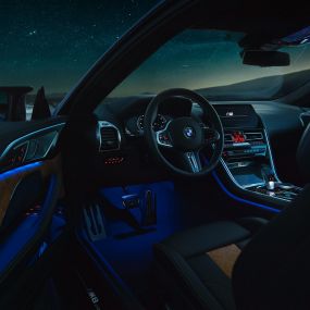 Интериор BMW M8 Competition Купе. BMW M8 Competition Купе, интериор през нощта с М кожен волан и синьо вътрешно осветление при отворена врата на водача.