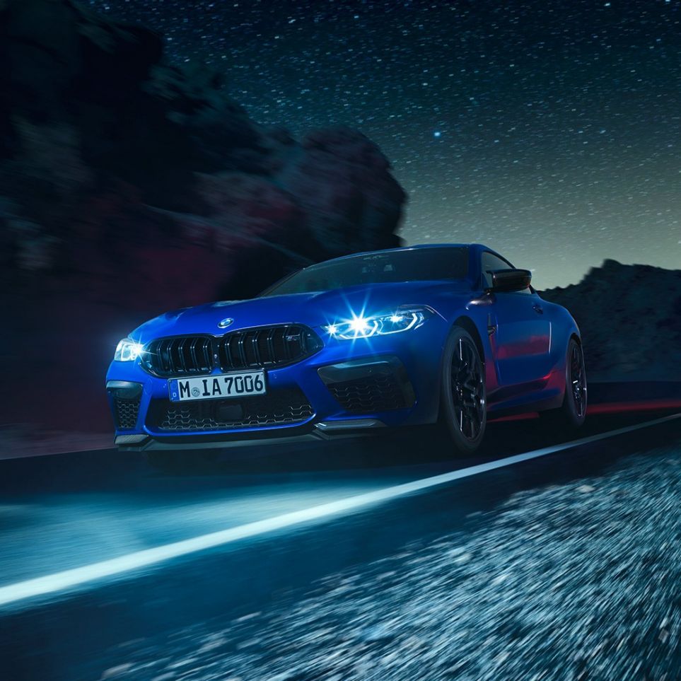 BMW M8 Competition Купе, три четвърти изглед отпред. BMW M8 Competition Купе в Marina Bay Blue metallic с ярки LED фарове, три четвърти изглед отпред в движение през нощта.