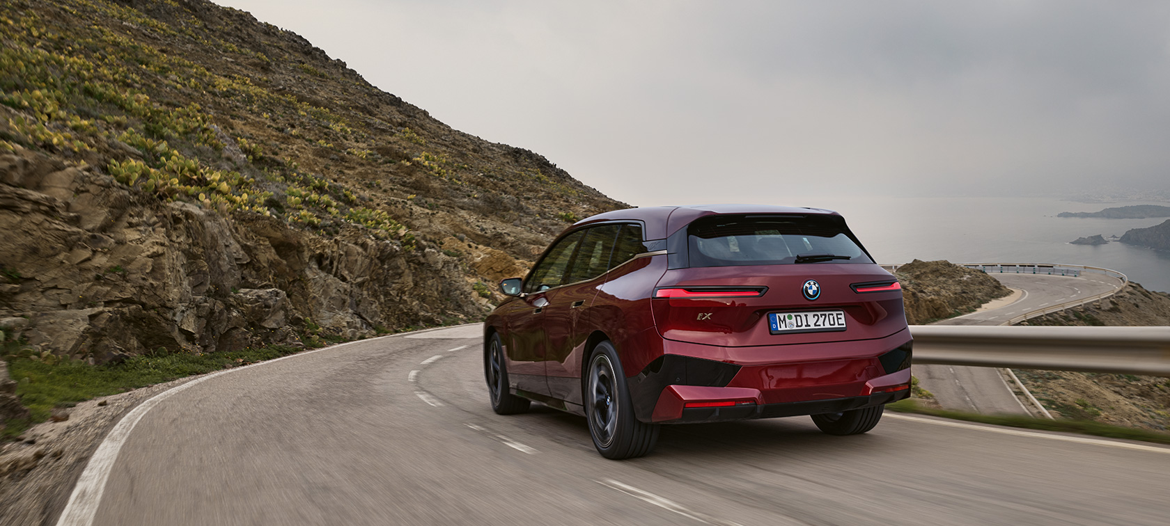 BMW iX i20 2021 електрически SUV BMW iX xDrive50 в движение по крайбрежен път BMW iX i20 2021 електрически SUV BMW iX xDrive50 Aventurine Red изглед отстрани в движение по крайбрежен път