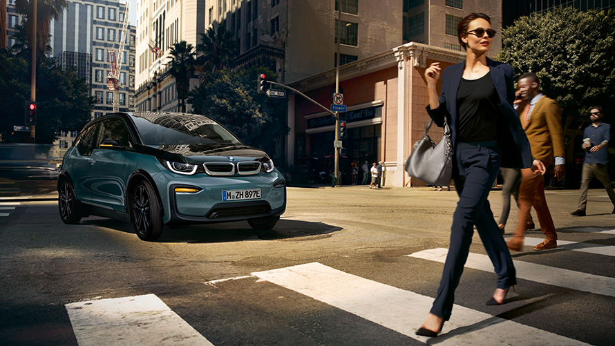 BMW I01 три четвърти изглед отпред с жена на пешеходна пътека BMW i3 I01 Blue Ridge Mountain metallic с акцент Frozen Grey metallic три четвърти изглед отпред с жена на пешеходна пътека