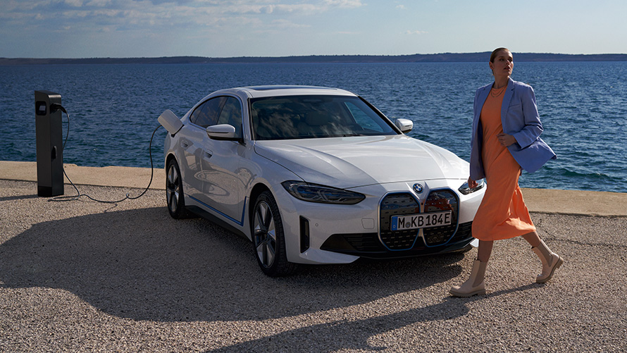 Три четвърти изглед отпред BMW i4 eDrive40 G26 2021 на зарядна станция Жена пред BMW i4 eDrive40 G26 2021 Mineral White три четвърти изглед отпред на зарядна станция пред езеро