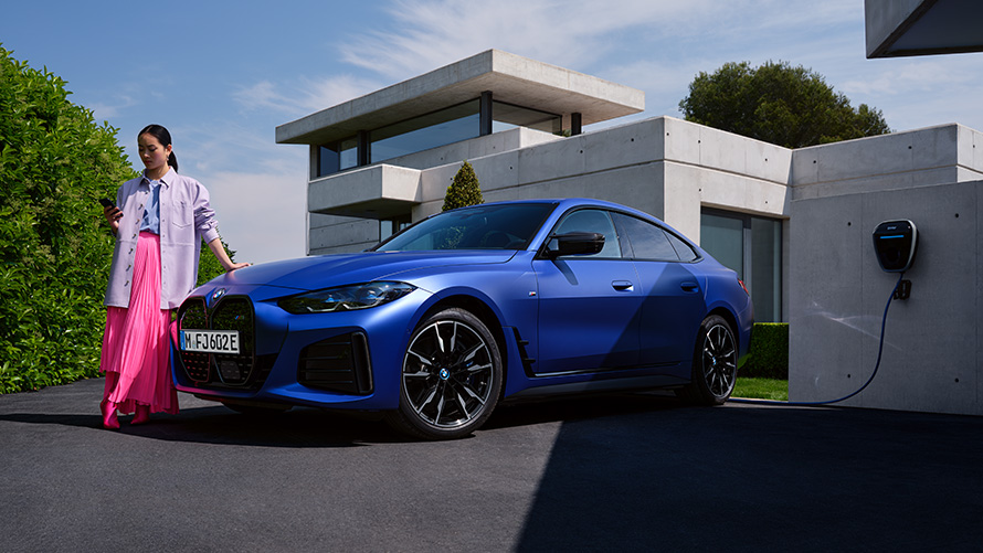 Три четвърти изглед отстрани BMW i4 M50 G26 2021 с жена пред къща BMW i4 M50 G26 2021 BMW Individual Frozen Portimao Blue metallic три четвърти изглед отстрани с жена пред къща