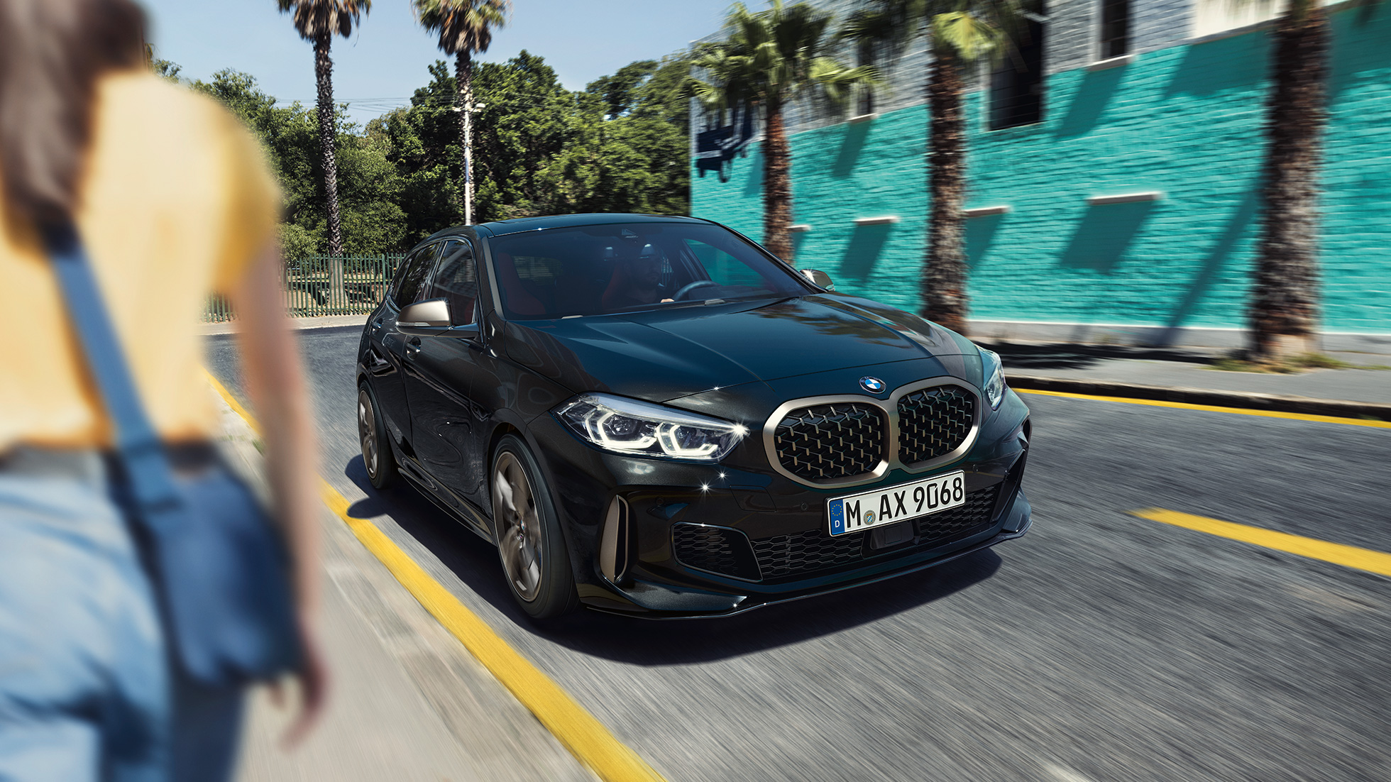 BMW M135i xDrive, три четвърти изглед отпред BMW M135i xDrive в Black Sapphire metallic, три четвърти изглед отпред в движение с жена на преден план