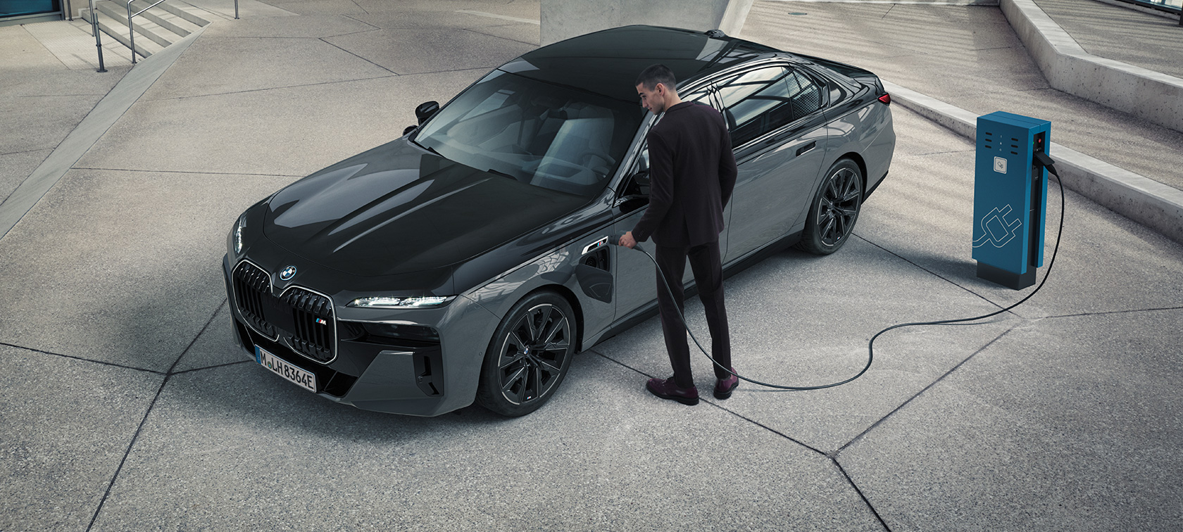 BMW M760e xDrive G70 M Performance пакет BMW M760e xDrive G70 M Performance пакет BMW BMW Individual Two-Tone лаково покритие Saphire Black metallic BMW Individual Dravite Grey metallic три четвърти изглед отпред с мъж със заряден кабел до зарядна станция