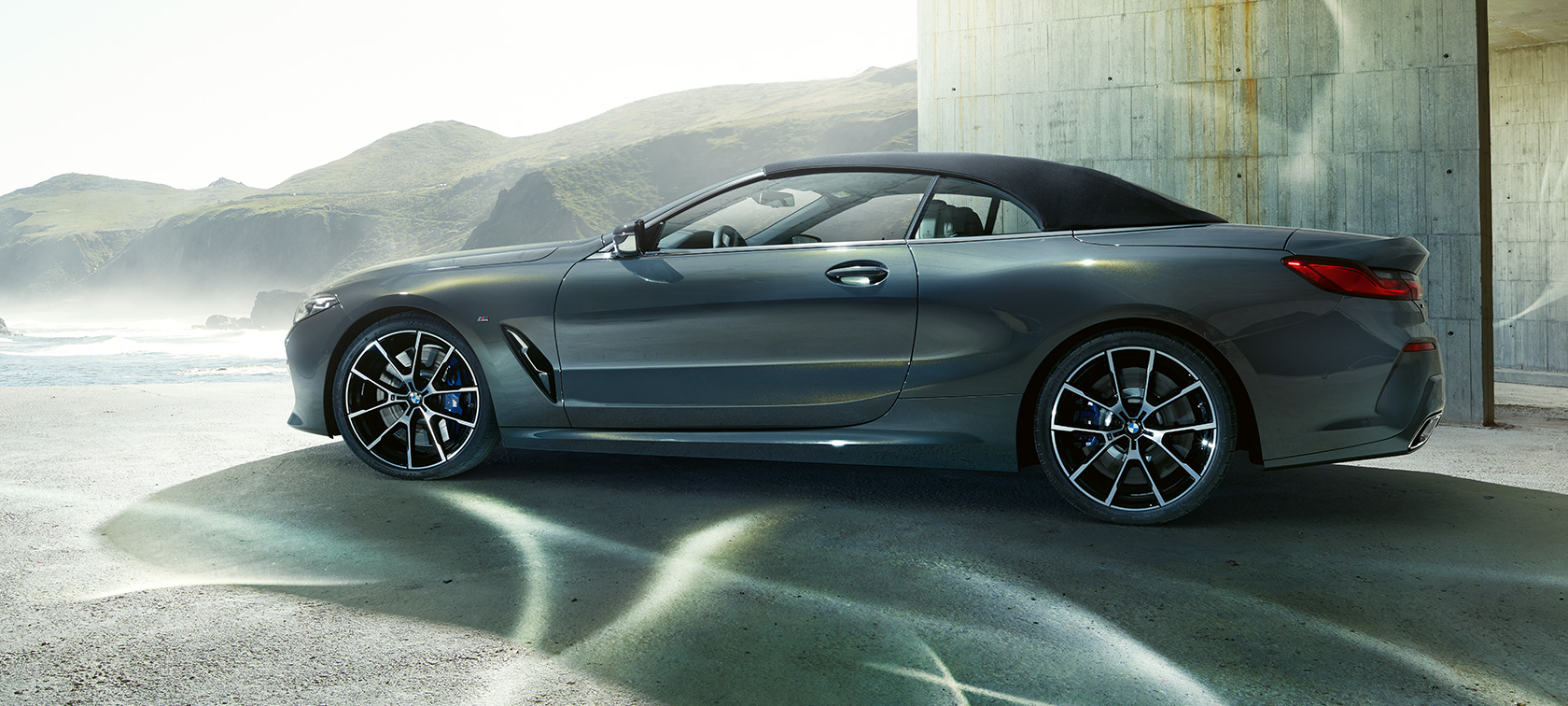 BMW M850i xDrive Кабриолет, изглед отстрани. BMW M850i xDrive Кабриолет в цвят Dravit Grey metallic със затворен мек покрив, изглед отстрани пред хълмове.