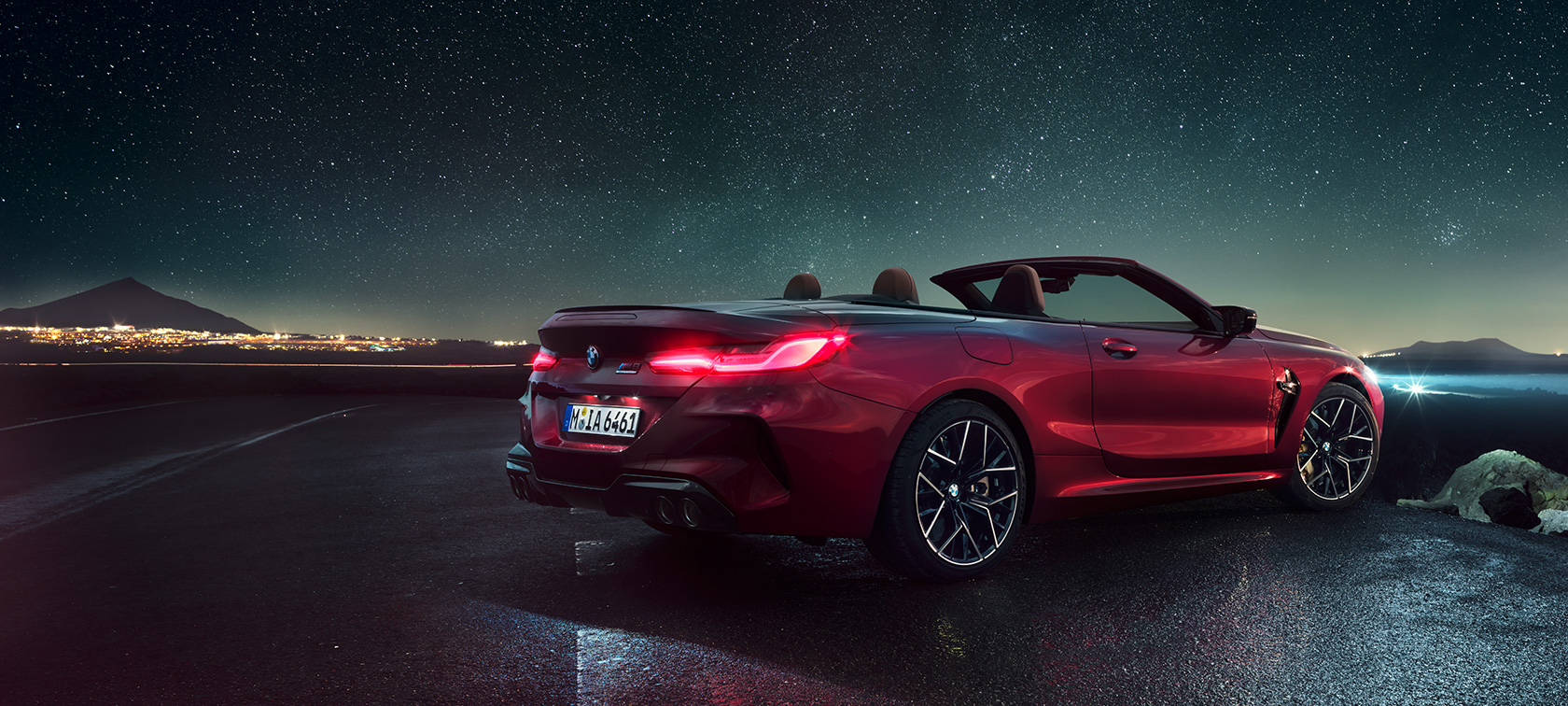 BMW M8 Competition, три четвърти изглед отзад. BMW M8 Competition в цвят Motegi Red metallic с отворен мек покрив, три четвърти изглед отзад, в покой под звездно небе.