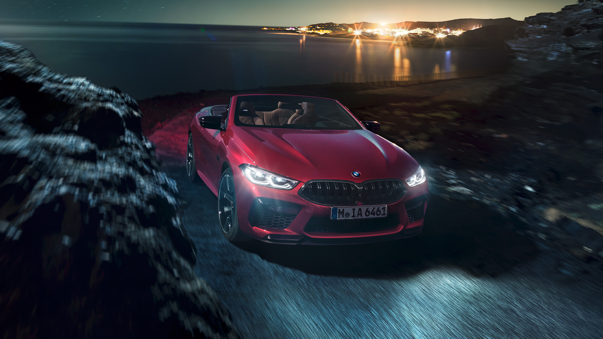 BMW M8 Competition Кабриолет в цвят Motegi Red metallic, три четвърти изглед отпред. BMW M8 Competition Кабриолет в цвят Motegi Red metallic с отворен мек покрив, три четвърти изглед отпред, в движение през нощта по крайбрежен път.