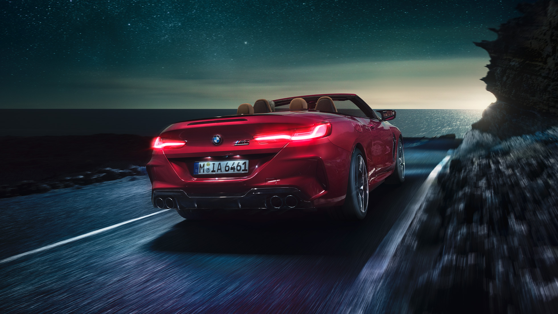 BMW M8 Competition Кабриолет в цвят Motegi Red metallic, три четвърти изглед отпред. BMW M8 Competition Кабриолет в цвят Motegi Red metallic с отворен мек покрив, три четвърти изглед отпред, в движение през нощта по крайбрежен път.
