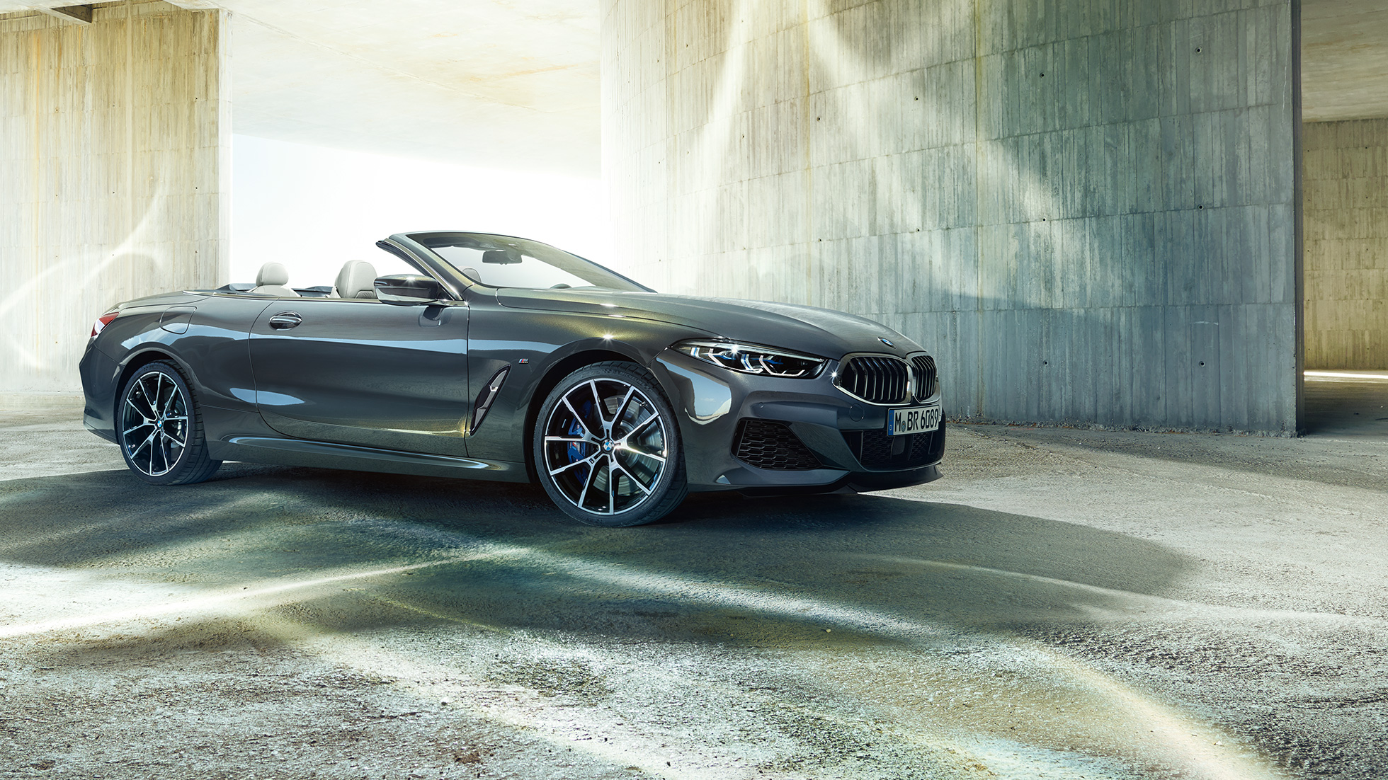 BMW M850i xDrive Кабриолет, три четвърти изглед отстрани. BMW M850i xDrive Кабриолет в цвят Dravit Grey metallic с отворен мек покрив, три четвърти изглед отстрани, в покой пред бетонна сграда.