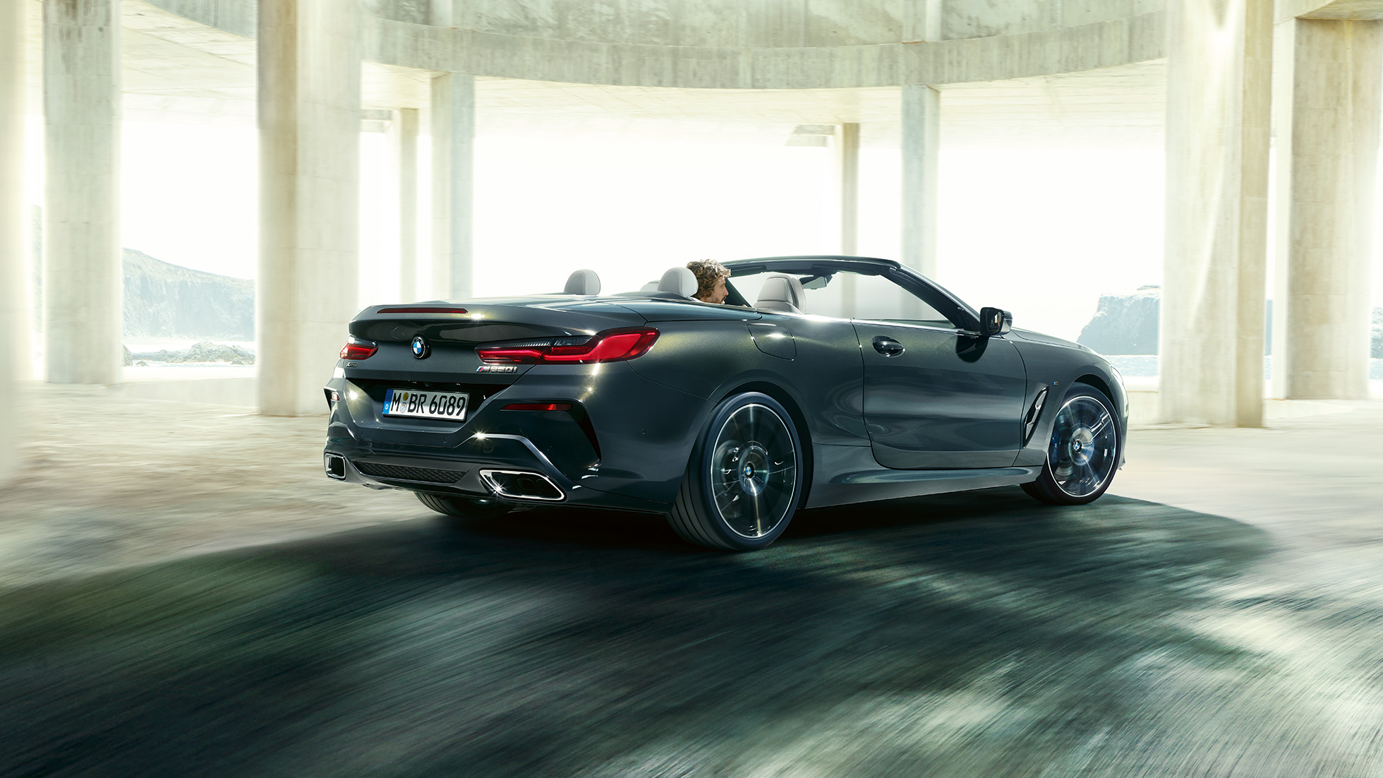 BMW M850i xDrive Кабриолет, три четвърти изглед отзад. BMW M850i xDrive Кабриолет в цвят Dravit Grey metallic с отворен мек покрив, три четвърти изглед отзад, в движение през бетонна сграда.