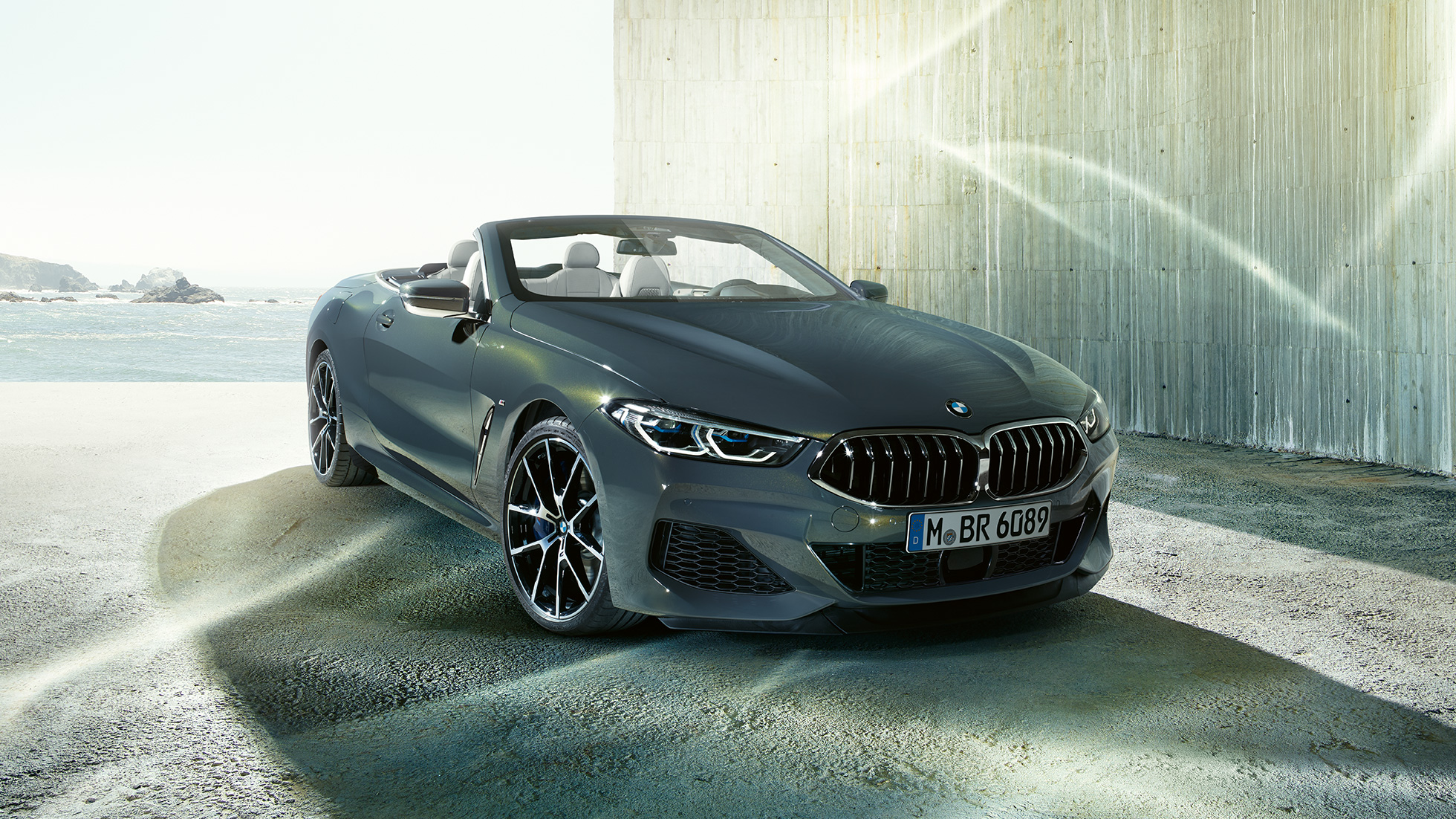 BMW M850i xDrive Кабриолет, три четвърти изглед отпред. BMW M850i xDrive Кабриолет в цвят Dravit Grey metallic с отворен мек покрив, три четвърти изглед отпред, в покой на брега.