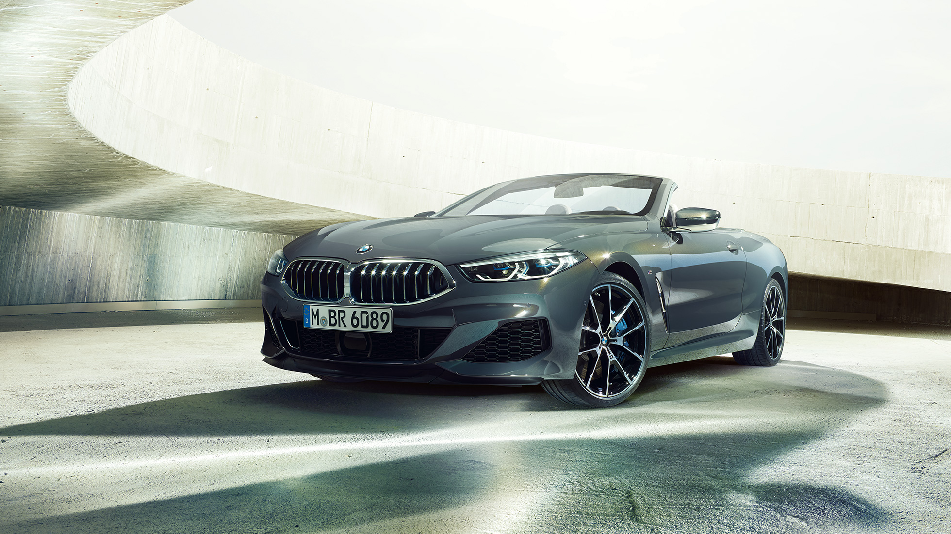 BMW M850i xDrive Кабриолет, три четвърти изглед отпред. BMW M850i xDrive Кабриолет в цвят Dravit Grey metallic с отворен мек покрив, три четвърти изглед отпред, в покой пред бетонна сграда.