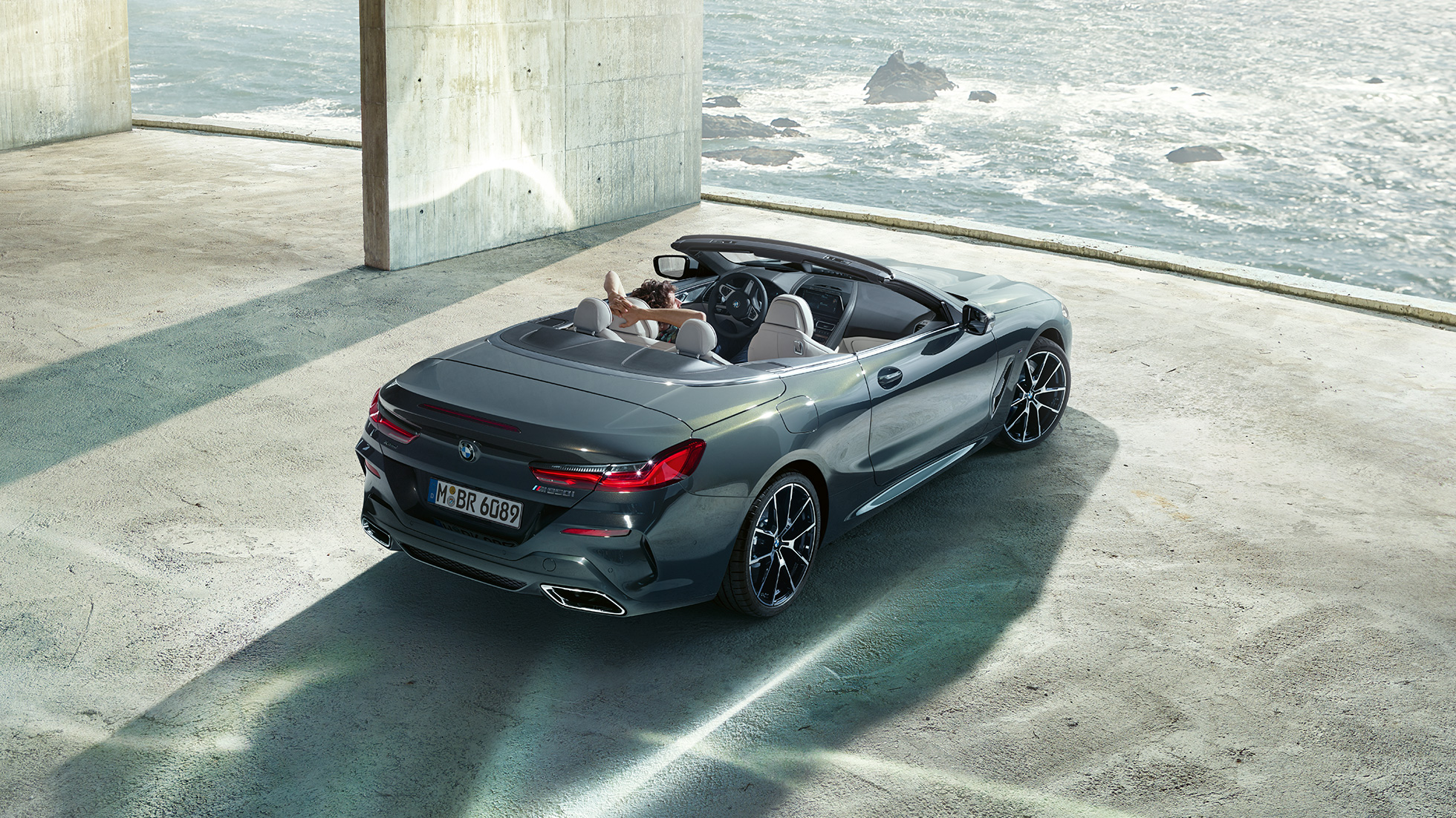 BMW M850i xDrive Кабриолет, от перспектива „птичи поглед“. BMW M850i xDrive Кабриолет в цвят Dravit Grey metallic с отворен мек покрив, от перспектива „птичи поглед“, паркирано в бетонна сграда на фона на океана.