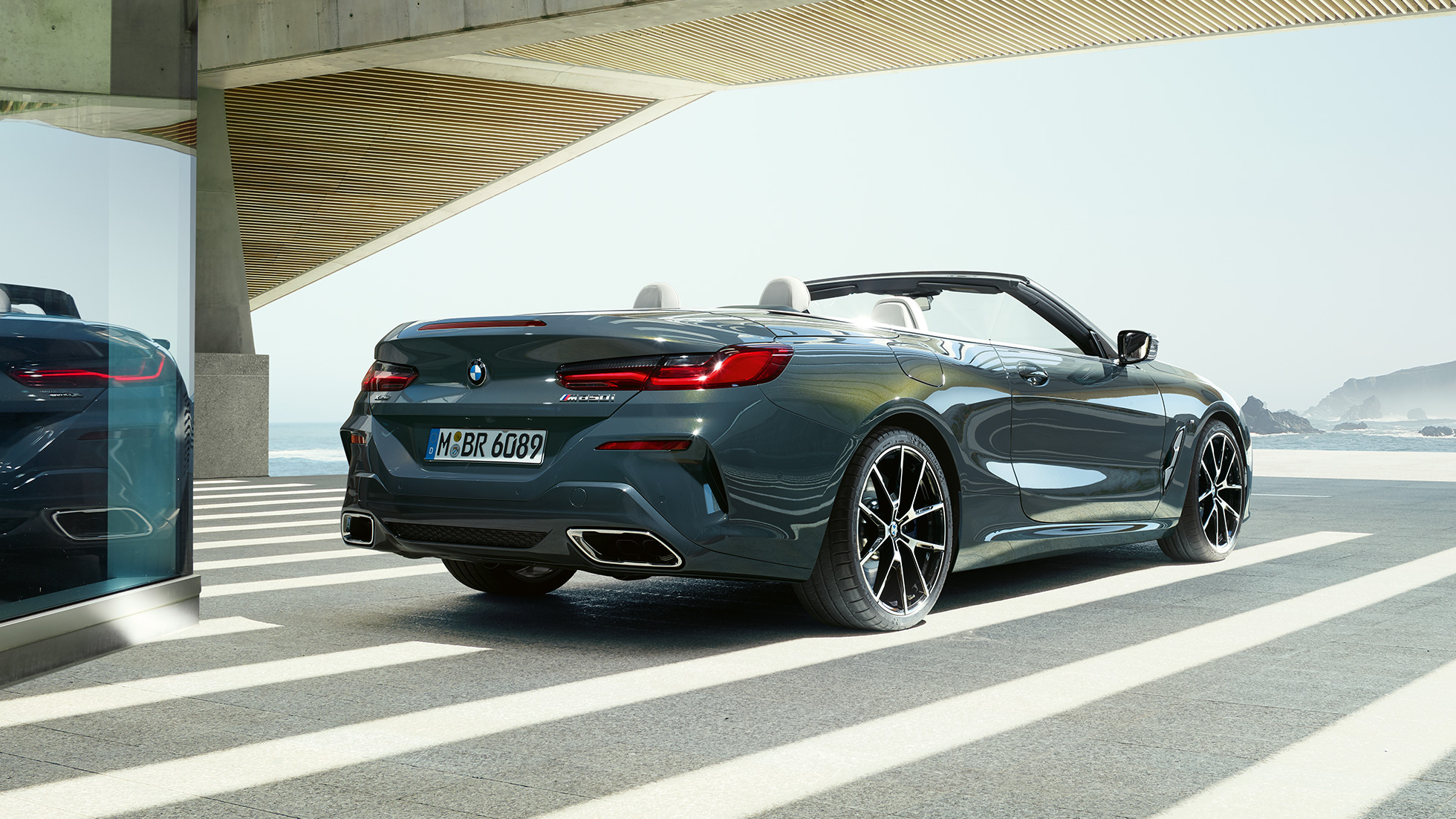 BMW M850i xDrive Кабриолет, три четвърти изглед отзад. BMW M850i xDrive Кабриолет в цвят Dravit Grey metallic с отворен мек покрив, три четвърти изглед отзад, паркирано с изглед към океана.