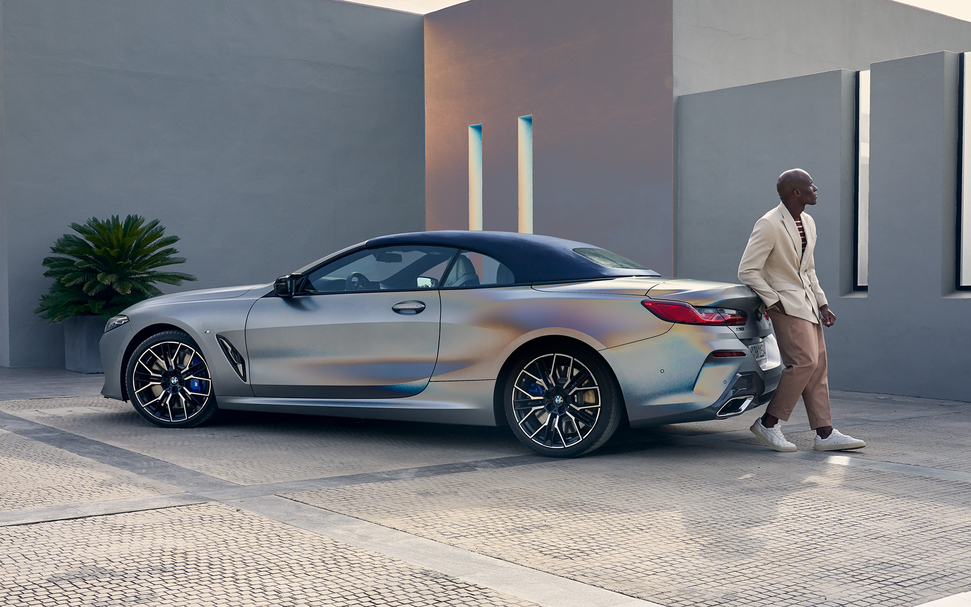 Три-четвърти изглед отзад BMW M850i xDrive Кабриолет G14 LCI Facelift 2022 BMW Серия 8 Кабриолет G14 LCI фейслифт 2022 BMW Individual Frozen Pure Grey metallic три-четвърти изглед отзад с мъж, облегнат на автомобила