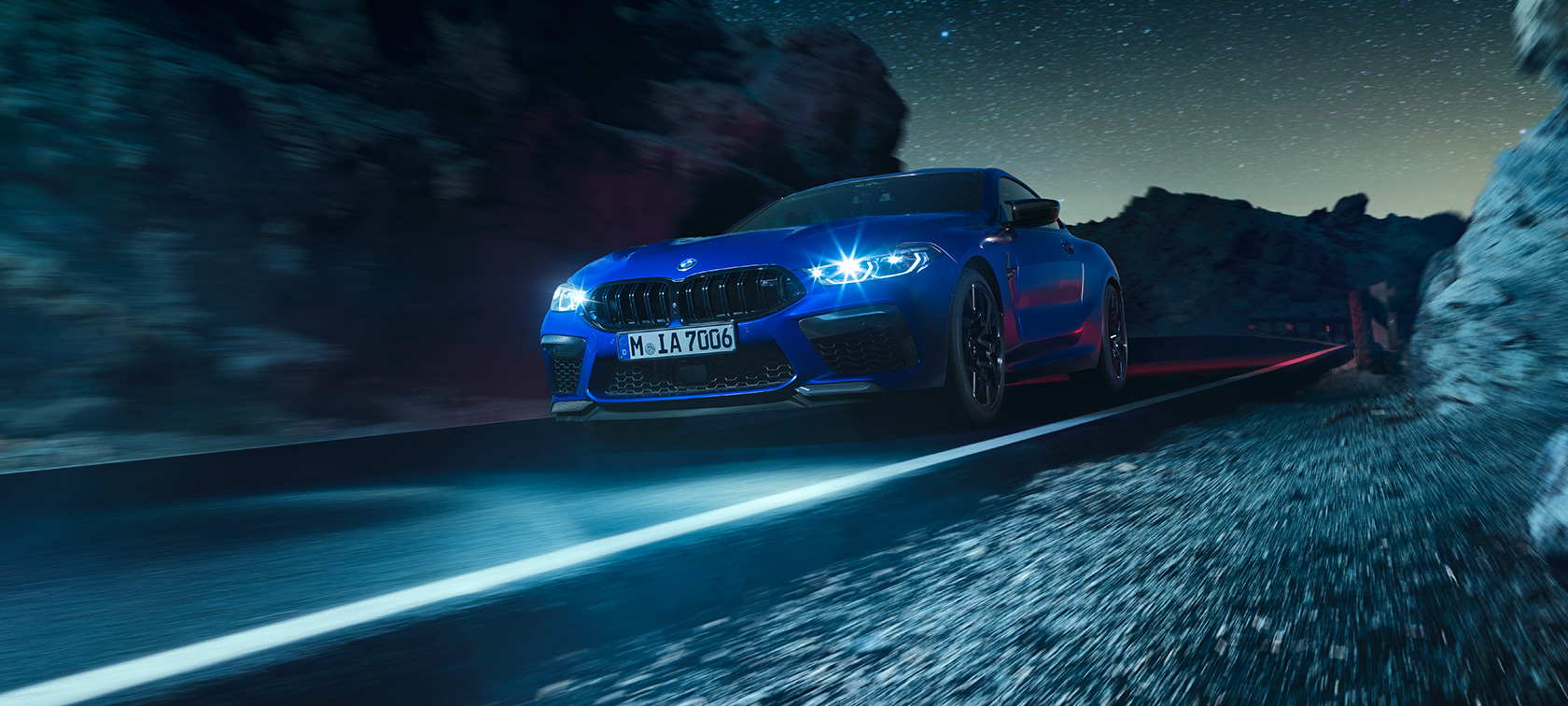 BMW M8 Competition Купе, три четвърти изглед отпред. BMW M8 Competition Купе в Marina Bay Blue metallic, три четвърти изглед отпред, в движение през нощта