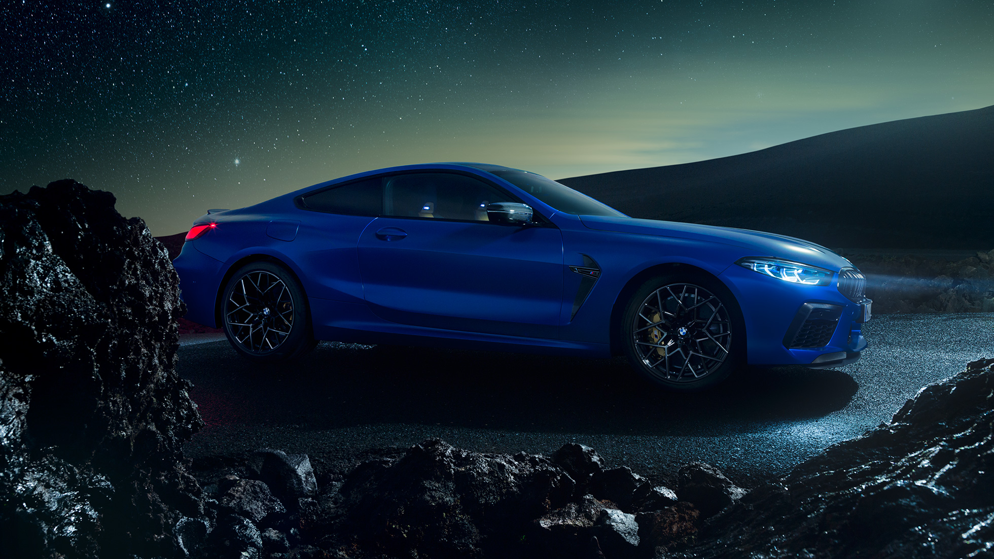 BMW M8 Competition Купе, три четвърти изглед отстрани. BMW M8 Competition Купе в Marina Bay Blue metallic, три четвърти изглед отстрани в покой през нощта.