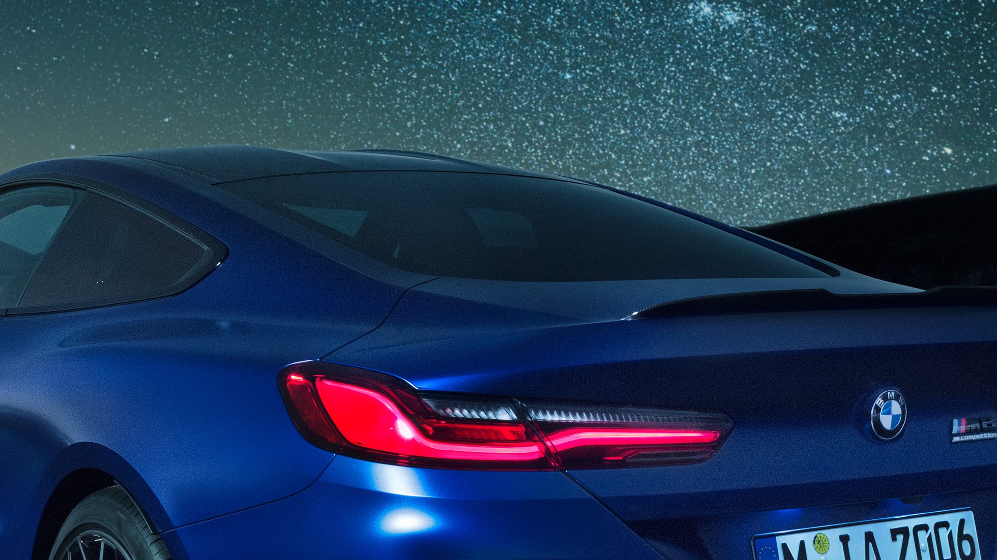 М Заден спойлер BMW M8 Competition Купе в Marina Bay Blue metallic, снимка отблизо на областта на раменете през нощта с М заден спойлер от карбон.