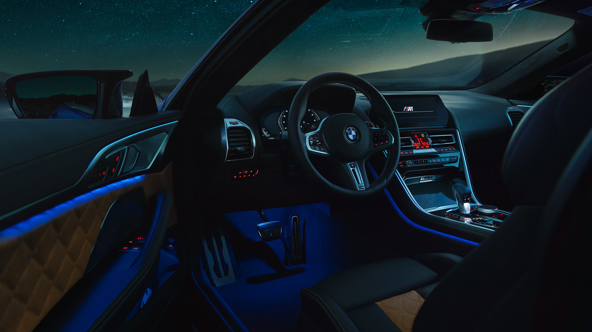 Интериор BMW M8 Competition Купе. BMW M8 Competition Купе, интериор през нощта с М кожен волан и синьо вътрешно осветление при отворена врата на водача.