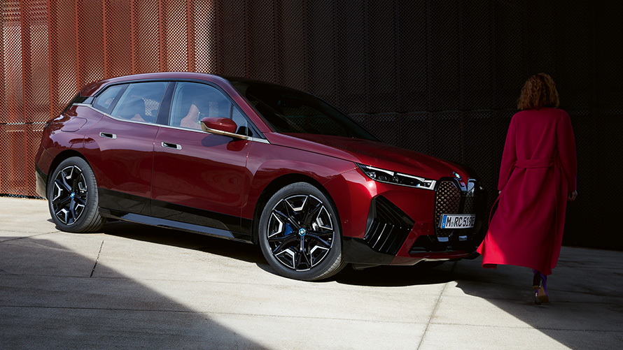 Жена пред BMW iX i20 2021 три четвърти изглед отпред Жена с червено палто пред BMW iX i20 2021 три четвърти изглед отпред