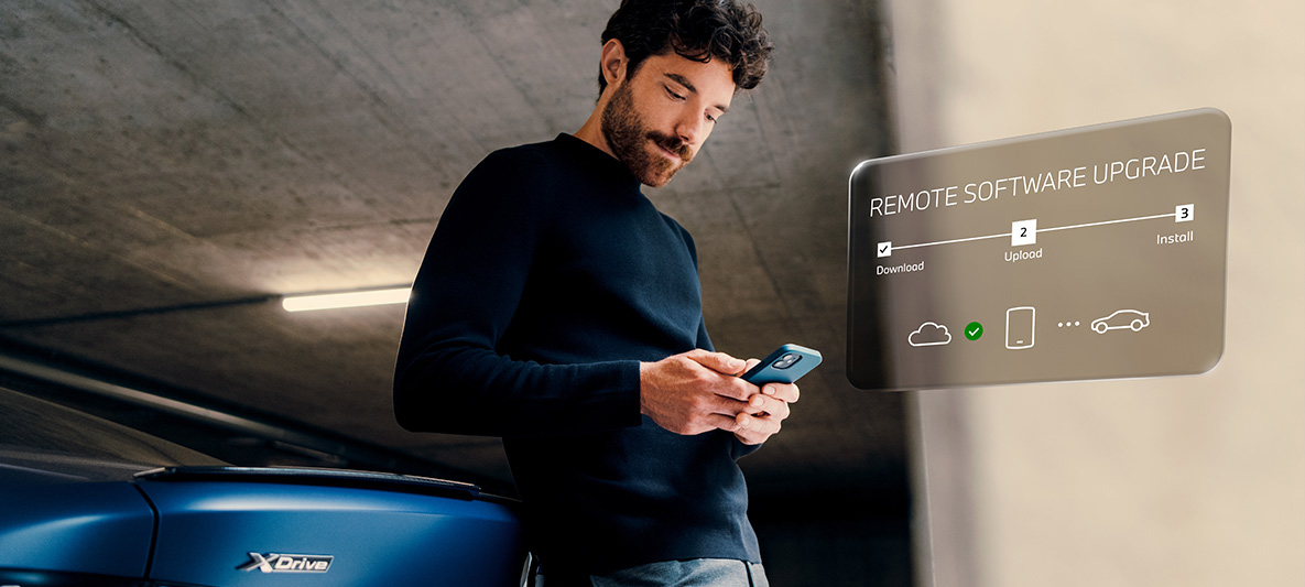 Мъж се обляга на задната част на BMW и гледа смартфона си Remote Software Upgrade мъж се обляга на BMW и гледа смартфона си