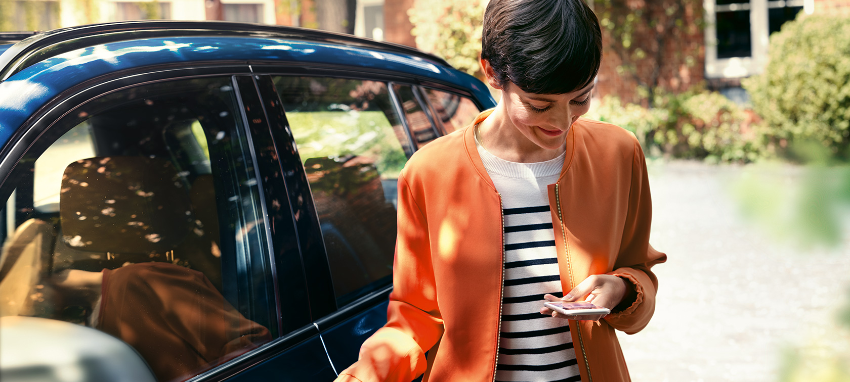Тъмнокоса жена пред BMW Тъмнокоса жена в оранжево яке пред BMW гледа смартфона си
