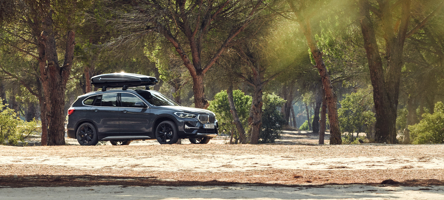 Страничен изглед на BMW X1 с BMW покривен багажник BMW летни оферти Страничен изглед на BMW X1 Mineral Grey с BMW покривен багажник, което е спряло в гора BMW летни оферти