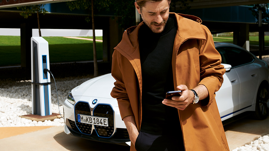 BMW Service Inclusive Продължителност и срокове за електрически автомобили BMW Service Inclusive мъж гледа смартфона си пред BMW i4 на зарядна станция