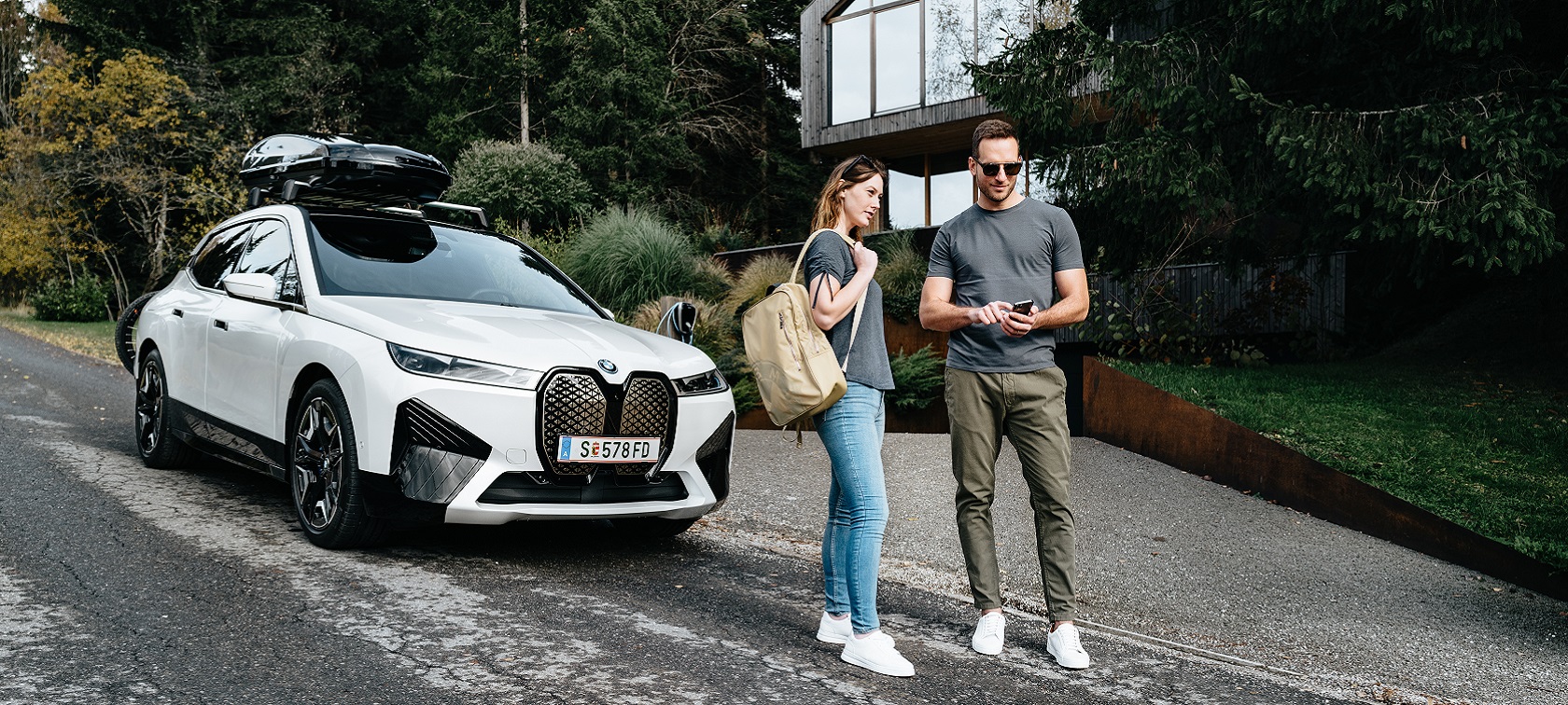 Мъж с телефон и жена с раница, стоящи пред BMW iX. Мъж с телефон и жена с раница, стоящи пред BMW iX.