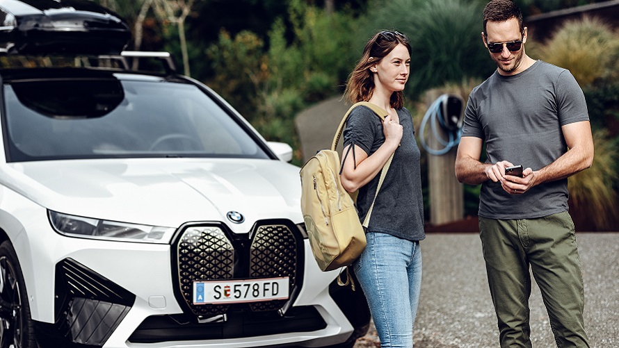 Мъж с телефон и жена с раница, стоящи пред BMW iX. Мъж с телефон и жена с раница, стоящи пред BMW iX.